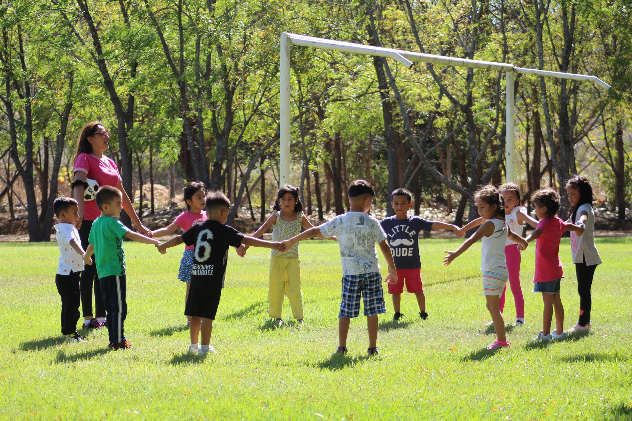 Kinder stehen mit erwachsener Frau auf einem Fußballplatz im Kreis und halten sich an den Händen