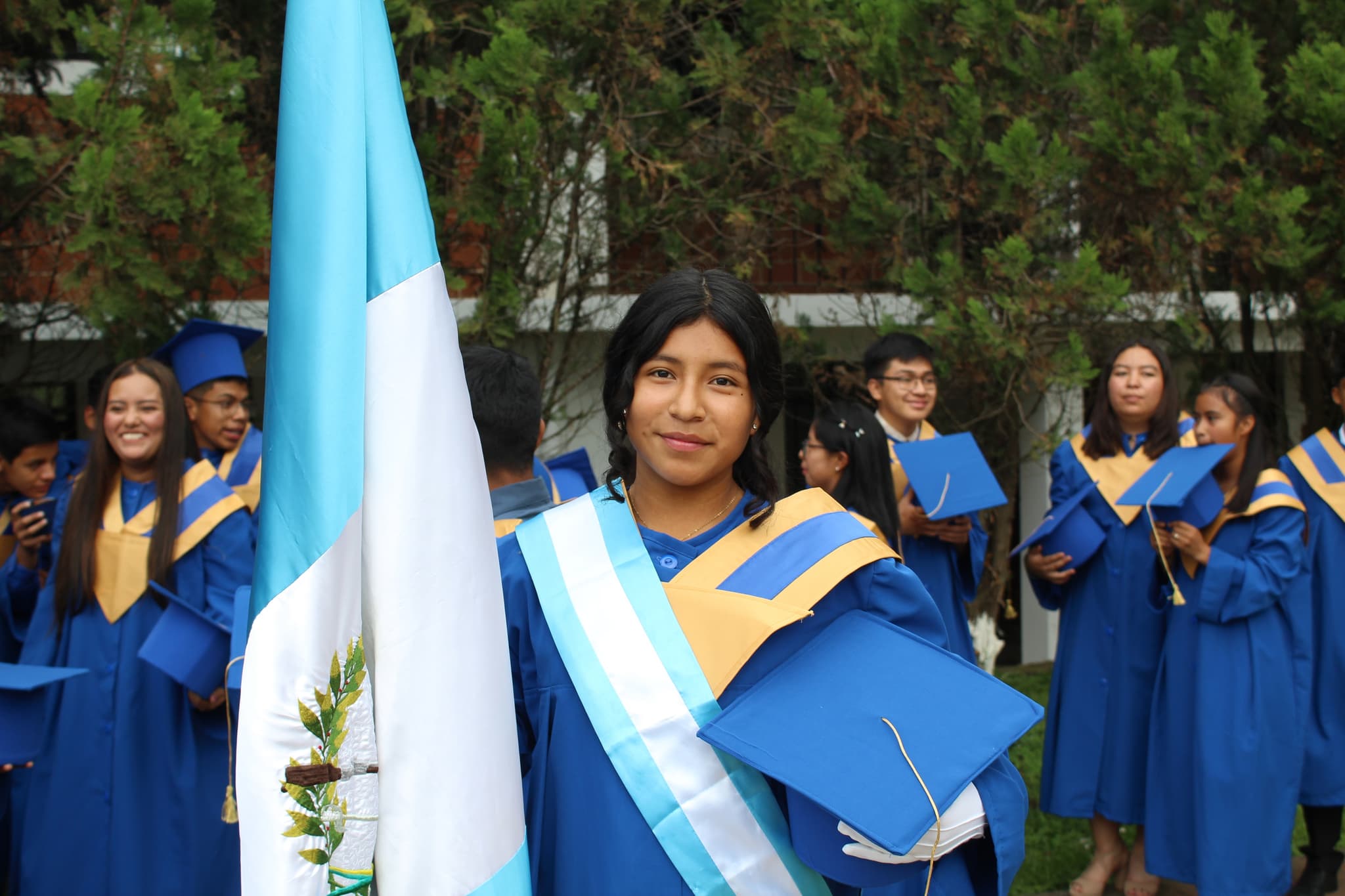 Ein Mädchen in einer blauen Absolventenuniform, die in einer Hand eine Guatemala-Flagge hält und in der anderen ihren Absolventenhut.