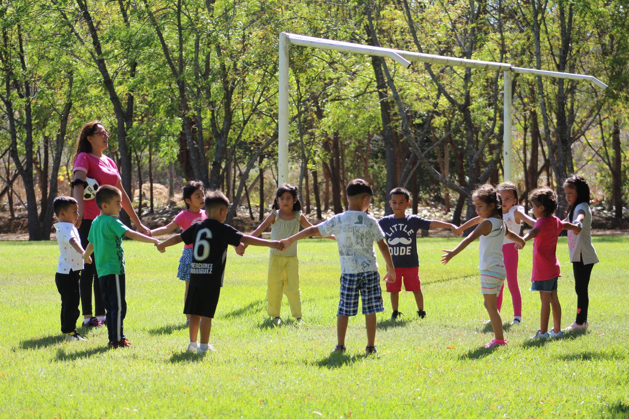 Kinder stehen mit erwachsener Frau auf einem Fußballplatz im Kreis und halten sich an den Händen