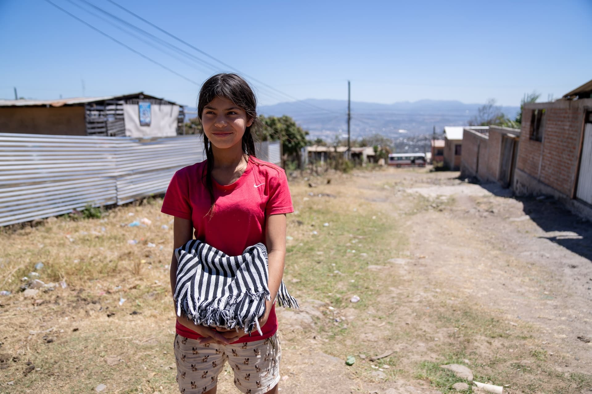 Mädchen mit einem roten Shirt, das mit einer Decke in der Hand auf einem verlassenen Gelände in Honduras steht.