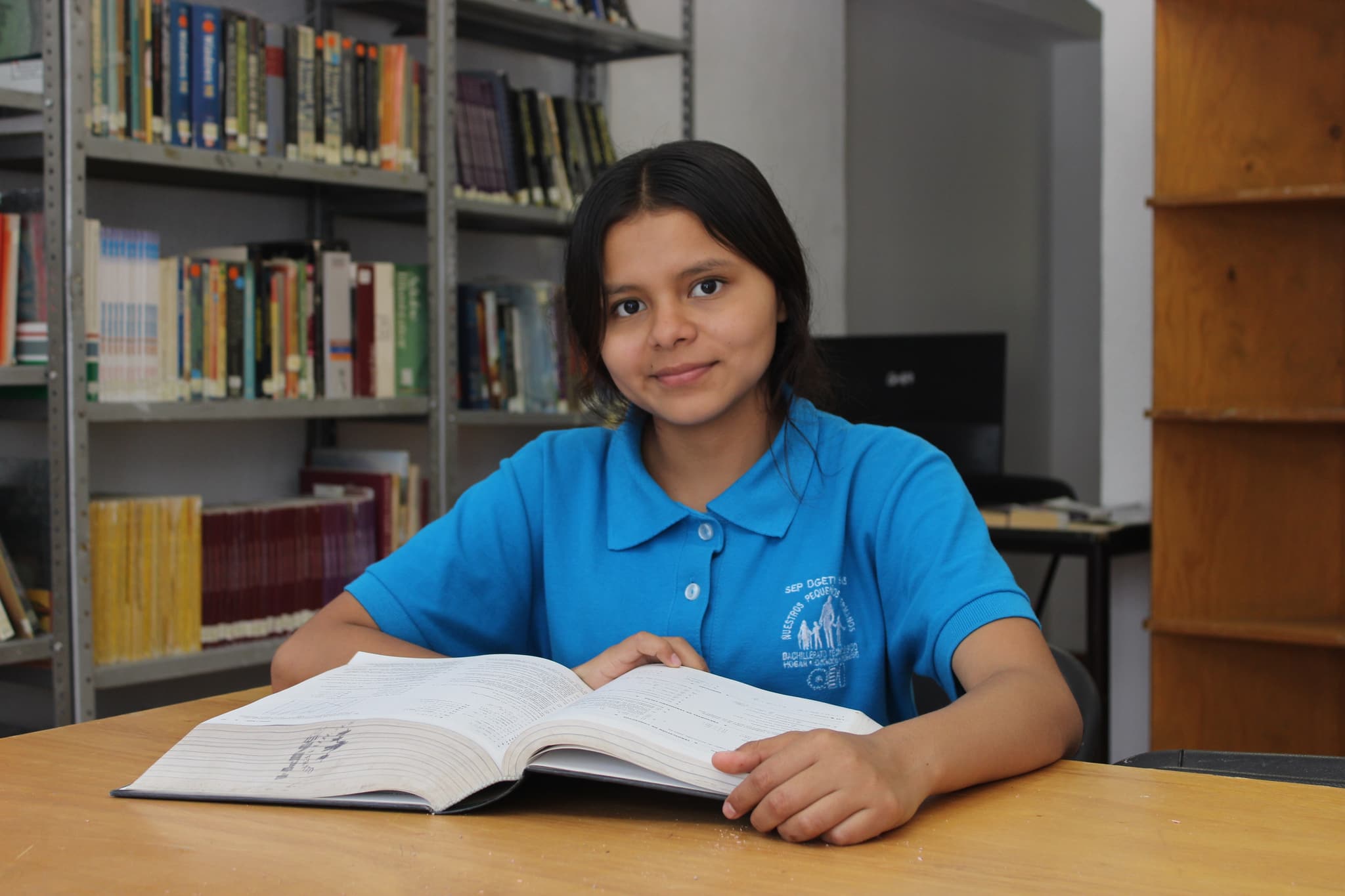 Mädchen mit einem nph-Shirt, das an einem Tisch mit einem Buch sitzt. Im Hintergrund sind große Bücherregale zu sehen.